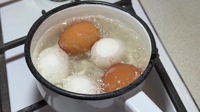 Close-up boiling eggs in enameled ladle on gas stove, brown and white chicken eggs simmering in bubbling water, steam rising, white