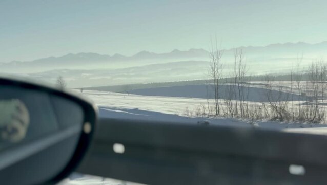 Soft winter light reveals distant misty mountains and wide snowy fields seen through a car side mirror creating a calm cinematic travel moment with gentle motion cold air and quiet atmospheric scenery