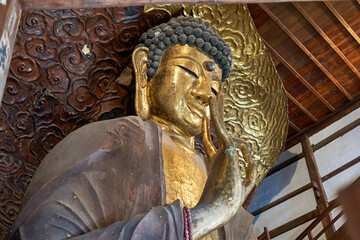 Obraz premium Close-up view of a large golden Buddha statue inside a traditional Japanese temple. The Buddha figure features a serene expression with closed eyes, elongated earlobes, and characteristic