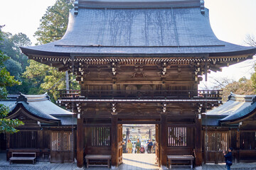 A traditional Japanese wooden temple gate (sanmon) with an imposing curved tiled roof stands as the central focus. The two-story structure features intricate dark timber joinery, ornate brackets, © Andrius