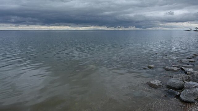 Video of a large lake with rocks along the shoreline. The water has small rippling waves reflecting a cloudy summer sky
