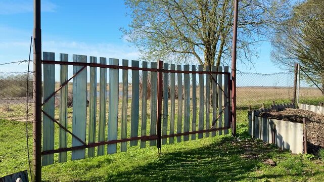 Weathered metal gate with vertical stainless strips, chain link fences flanking sides, spring grass carpeting ground, clear blue