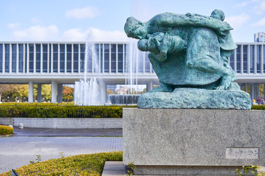 A bronze statue with green patina depicting a mother carrying two children, positioned on a granite pedestal with Japanese inscription reading "Mother and Child in the Storm." The sculpture shows
