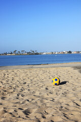 Obraz premium Blue and yellow soccer ball sits on soft Sail Bay sand, overlooking calm blue water, palm trees, and shoreline homes beneath a bright, cloudless San Diego sky, Southern California summer; copy space