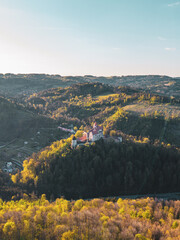 Fototapeta premium Pernstejn castle sits on a hill surrounded by trees. The sun shines over the landscape. Rolling hills and green forests fill the view. The area is peaceful and untouched.