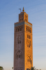 Fototapeta premium The Kutubiyya Mosque tower stands tall in Marrakesh. The sun sets in the background, casting a warm glow on the mosque's structure. Visitors admire the view as day ends.