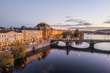 Fototapeta premium Aerial view of the National Theatre in Prague during the evening. The building is illuminated and reflects in the river. Bridges and streets show city life along the embankment.