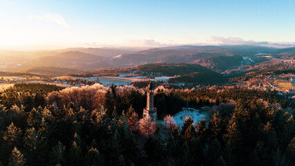 Stepanka lookout tower stands tall in the Jizera Mountains during winter. The sun sets behind distant hills, casting light over frosty trees and the surrounding landscape. © pyty