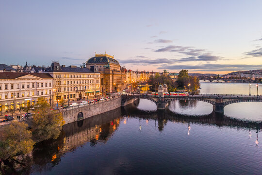 Aerial view of the National Theatre in Prague during the evening. The building is illuminated and reflects in the river. Bridges and streets show city life along the embankment.