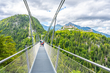 Fototapeta premium Visitors walk on Highline 179, a suspension bridge connecting castle ruins and a Roman fortress over a river gorge. Lush mountains surround the area, creating a scenic backdrop.