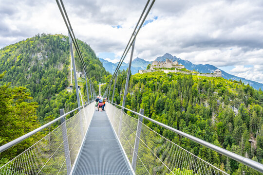Visitors walk on Highline 179, a suspension bridge connecting castle ruins and a Roman fortress over a river gorge. Lush mountains surround the area, creating a scenic backdrop.