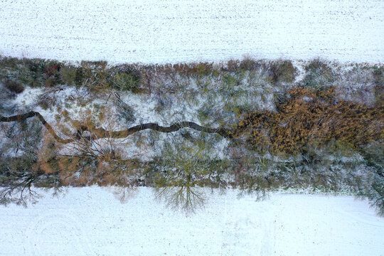 Aerial view of a winding stream flowing through a row of trees and shrubs amidst snow-covered fields Forchheim, Bavaria, Germany.