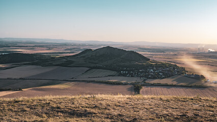 Fototapeta premium Rana Hill rises above the fields and village in Bohemian Central Mountains during sunset. The landscape shows varying colors of earth and sky. Smoke rises from distant factories.
