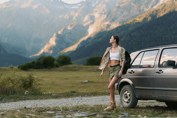 Fototapeta premium Woman stands beside a rugged car in a mountain meadow, wearing shorts and a cropped top with a jacket, embracing an adventurous outdoor lifestyle and wide horizon, wind in hair, sense of freedom,
