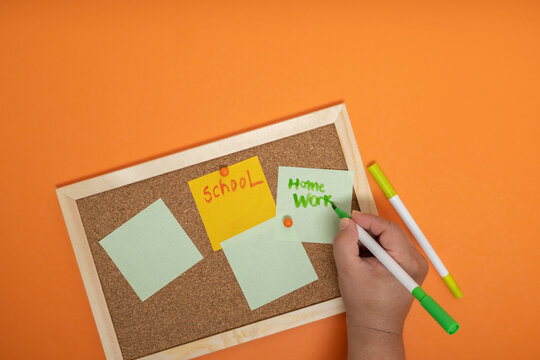 Close-up of hand writing homework reminder on sticky notes pinned on cork board, concept of planning, tasks and productivity.