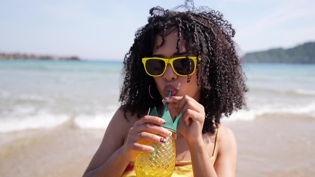 Beautiful mixed race woman with curly hair and yellow sunglasses smiling and drinking a refreshing fruit cocktail from a pineapple shaped cup on a sunny day at the seaside, enjoying her vacation