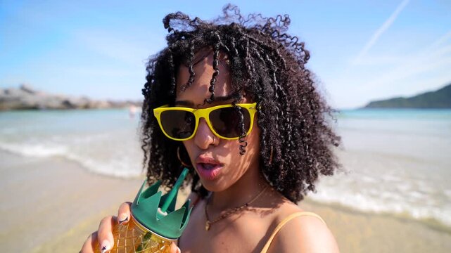 Attractive young woman with curly hair filming herself while enjoying a refreshing pineapple shaped drink at the beach on a sunny day, smiling and looking at the camera during her vacation