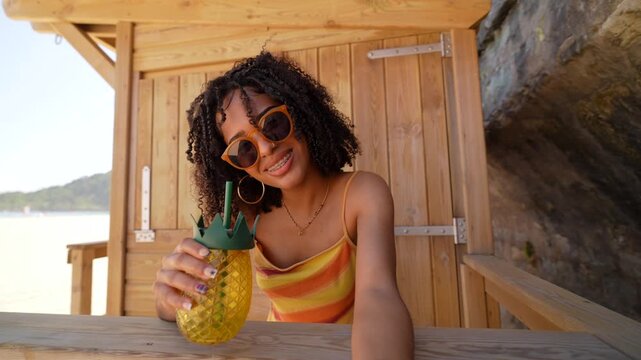 Beautiful young woman with curly hair and sunglasses taking a selfie video at a beach bar, smiling at the camera and holding a refreshing pineapple shaped drink on a sunny summer day