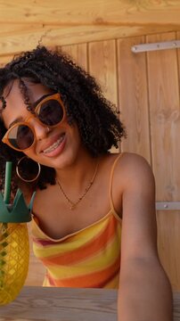 Cheerful young woman with curly hair and sunglasses smiling at the camera while enjoying a refreshing drink from a pineapple shaped cup at a beach bar on a sunny summer day