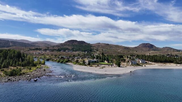 Aerial view of a river flowing into the deep blue waters of Nahuel Huapi Lake in Patagonia, Argentina. Small settlements sit on the rocky shoreline against a dry, rugged mountain backdrop.