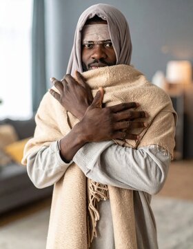 portrait of a young african american man feeling cold and shivering wrapped in a warm beige blanket, health recovery, flu symptoms, winter season home comfort