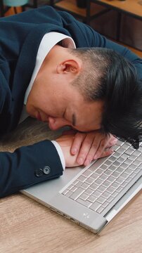 Bored sleepy Asian businessman worker working on laptop computer, leaning on hand falling asleep at office desk. Exhausted tired freelancer workaholic middle-aged man. Employment, occupation, workless