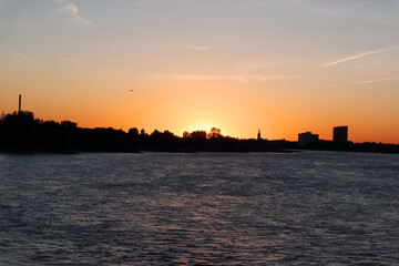 Fototapeta premium Sunset paints the sky in warm orange. Water glimmers with reflections of the fading light. Silhouetted trees and buildings form a distant skyline. This tranquil scene captures the quiet end of a day