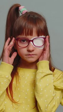 Sick ill preteen girl looking at camera having protective bandage plaster patch scratch on one eye. Injury trauma blind. Child kid wearing glasses at gray background. Medical eyesight vision therapy.
