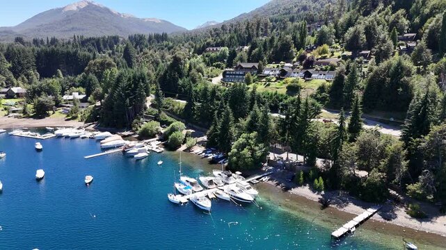 Stunning aerial perspective of numerous white boats and yachts moored in a tranquil blue bay of Nahuel Huapi Lake in Patagonia, Argentina. Lush alpine pine forests cover the rolling mountainside.