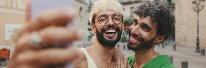Gay couple stands hugging and makes video call from mobile phone, waving in greeting. Panoramic © Andrii Nekrasov