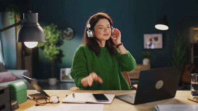 A woman sits at her desk wearing headphones. She is using her phone and has a notebook opened. A laptop is in front of her. The room has green walls and plants.