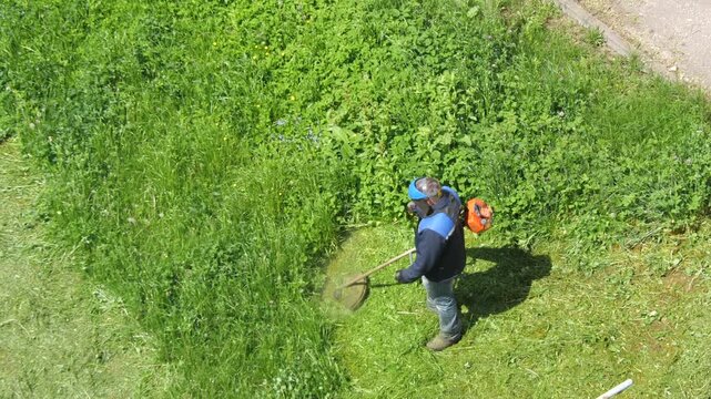 Gardener wearing safety equipment performing professional lawn care and landscape maintenance, cutting long grass and weeds with a petrol brush cutter on a sunny day