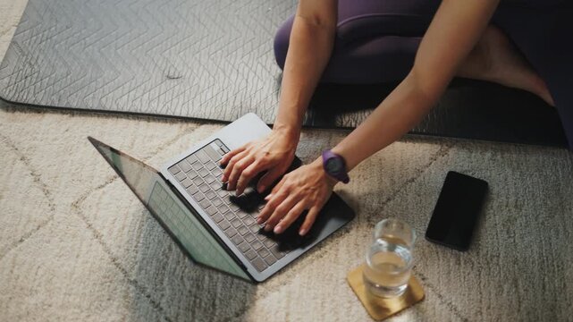 A person is sitting on a mat and working on a laptop. There is a glass of water and a phone nearby. The focus is on the laptop and the person's hand typing.