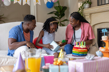 Fototapeta premium African American family celebrating on sofa at home, child opening gift box with cake