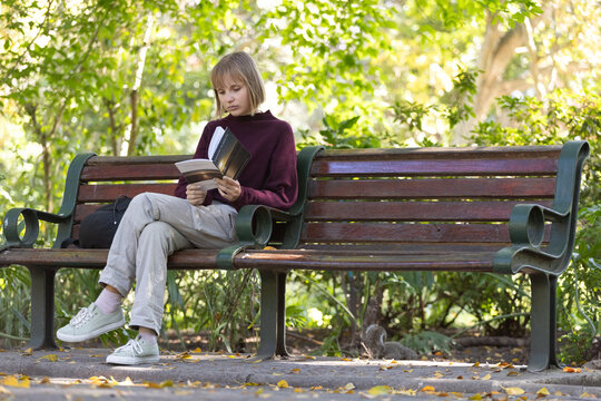 Woman in twenties sitting on left of wooden park bench reading paperback book with backpack