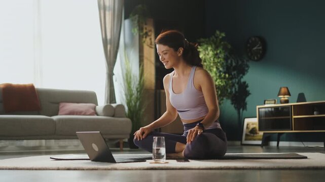 A woman practices yoga in a bright room at home. She sits on a mat with a laptop in front of her. A glass of water is nearby on a small table while she focuses on her session.
