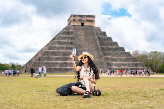 Young smiling Latina woman happily taking a selfie, expressing joy excitement while visiting Kukulkan pyramid at Chichen Itza, Yucatan Mexico, wearing summer outfit with hat, sunglasses in sunny day