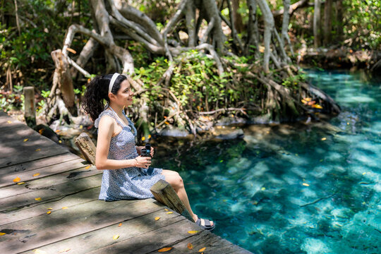 Young woman stands relaxed and smiling enjoying peaceful in Celestun Yucatan, surrounded by lush forest and clear lagoon, wearing light floral dress, sandals and casual accessories in tropical setting