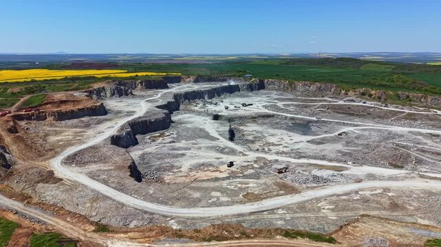 A large quarry site shows heavy machinery working on rocky terrain. Green fields stretch in the distance under a clear blue sky. Dust is present from ongoing activities.