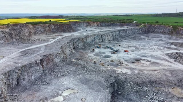 A large gravel quarry is active with machinery and trucks working. The surrounding land is green with fields and hills in the distance. The sky is clear and the sun is shining.