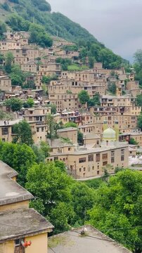 Panoramic view of historic Masouleh terraced mountain village showing traditional clay houses and a shrine with a green dome, panning left to right, vertical 9:16 video.