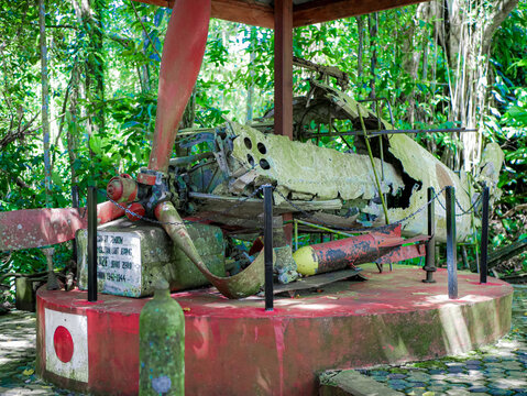 The moss-covered wreckage of a Japanese Zero fighter plane is displayed in an open-air monument against a backdrop of green forest.