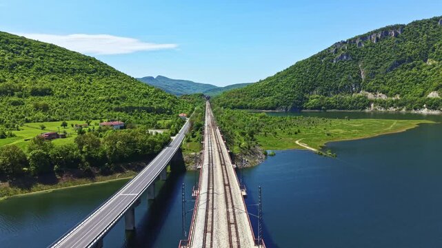 Train tracks stretch into the distance beside a river with green hills on both sides. A highway runs parallel to the tracks. The scene shows clear skies and bright daylight.