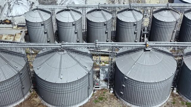 Storage silos stand in rows at a grain facility. Machinery is visible nearby, and the scene is bright with clear weather. The tanks are large and cylindrical, used for grain storage.