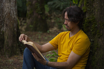 Obraz premium Side view of an adult man leaning against a tree trunk while reading a book outdoors.