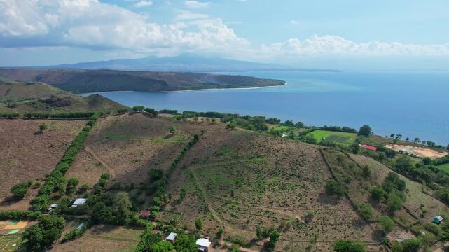Aerial drone footage of coastal hills in Kilo city area, Bima regency, Sumbawa island, Indonesia, with red onion fields around, blue quiet sea, and a large bay at the horizon with coral reef