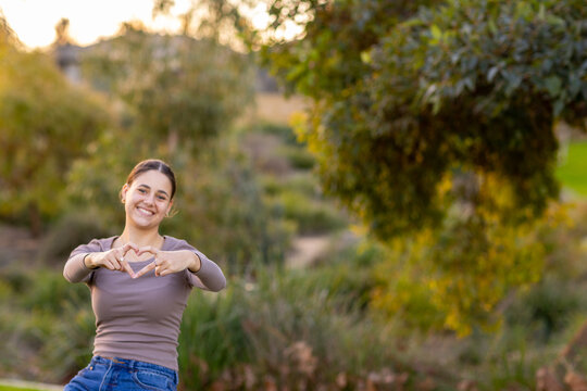 smiling girl holding fingers in heart shape with blurred background of foliage