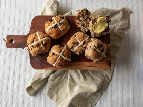 Whole and cut hot cross buns with butter on wooden serving tray with white table cloth, linen napkin