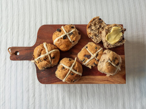Whole and cut Hot Cross Buns with butter on wooden serving board on white table cloth