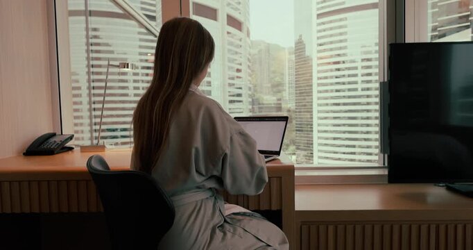 Woman in a robe typing on a laptop at a desk in a modern hotel room overlooking urban skyscrapers during the day. Slow zoom in camera movement from behind.